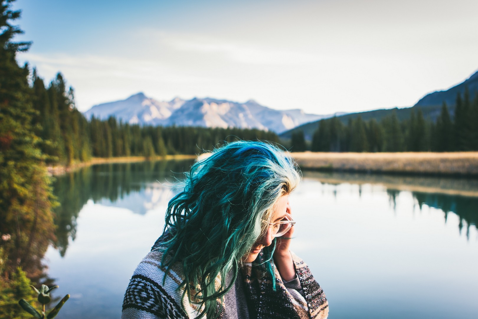 Mulher olhando para baixo, de perfil, cabelos azuis com mesclas loiras, em uma paisagem repleta de coníferas, com um lago atrás e montanhas ao fundo.